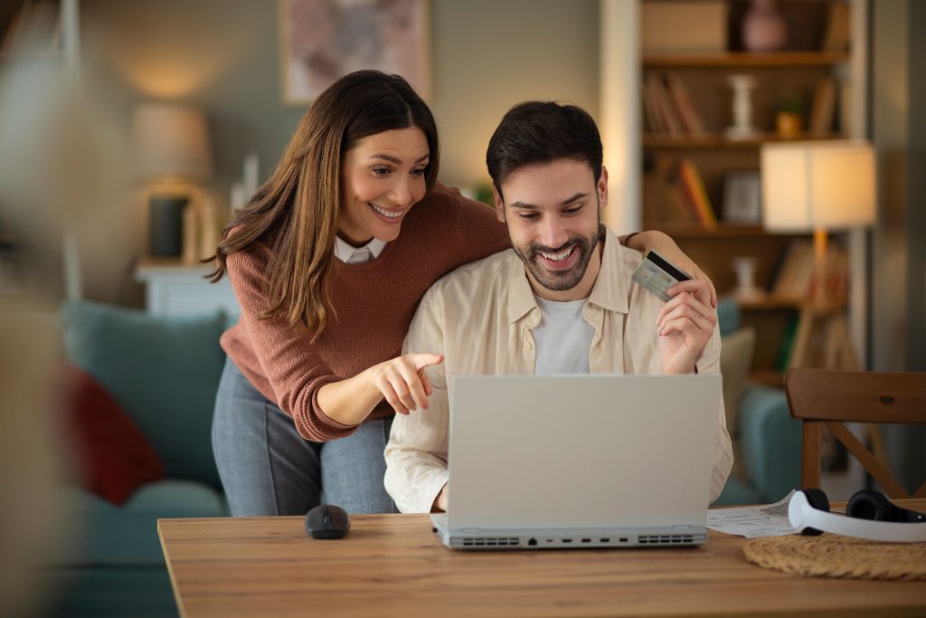 Young couple at home kitchen table looking at a computer. A man is holding a credit card and a woman is pointing at the computer screen.