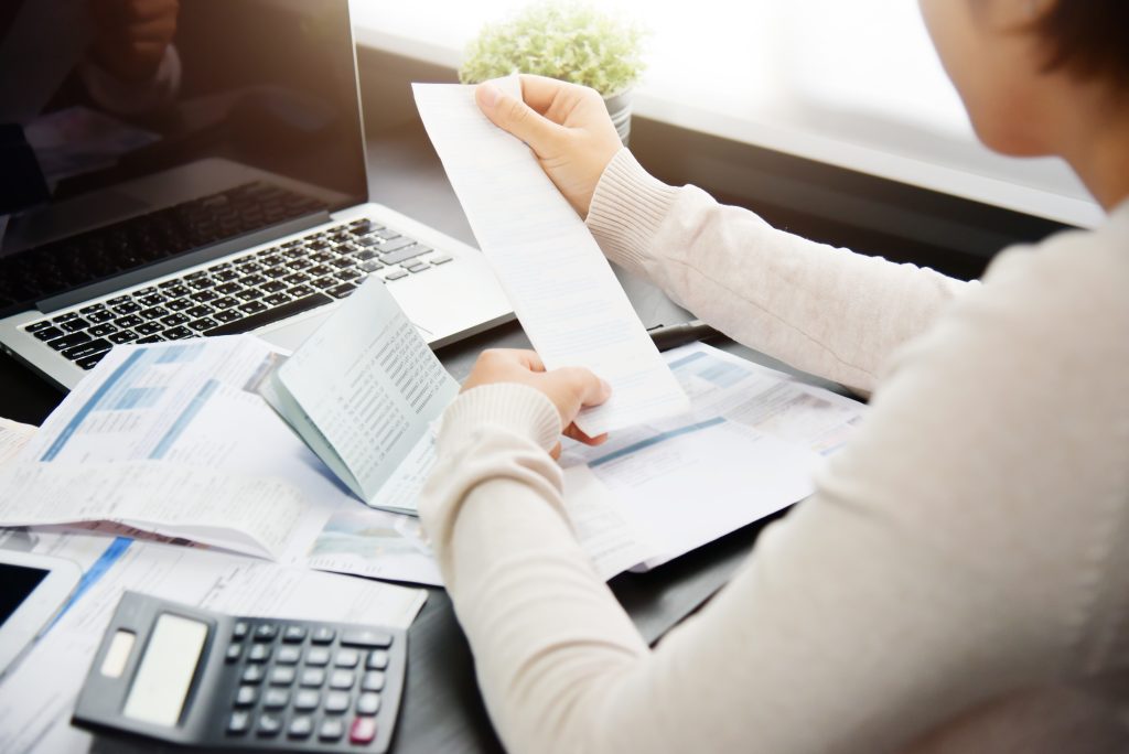 Woman sitting at a desk holding up a receipt with a computer, calculator, and papers on the desk. 