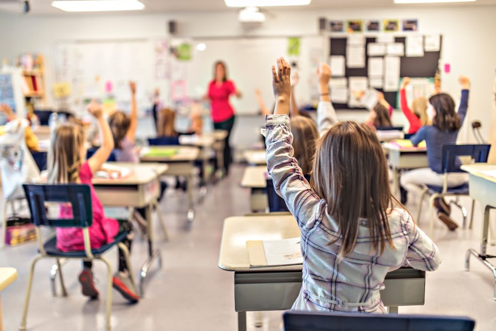 Group of students in a classroom raising their hands to answer teacher's question. 