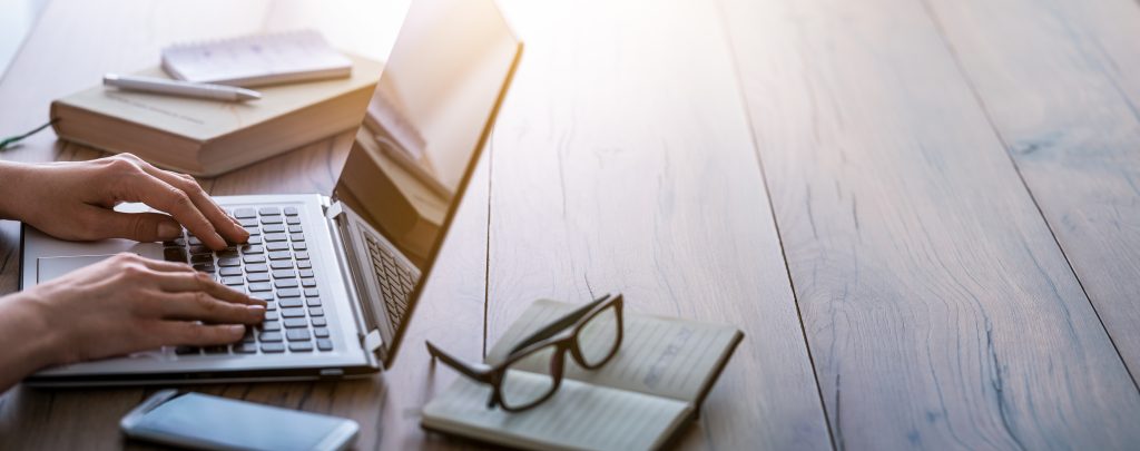 Someone typing on a computer at a desk with a notebook and glasses on the desk. 