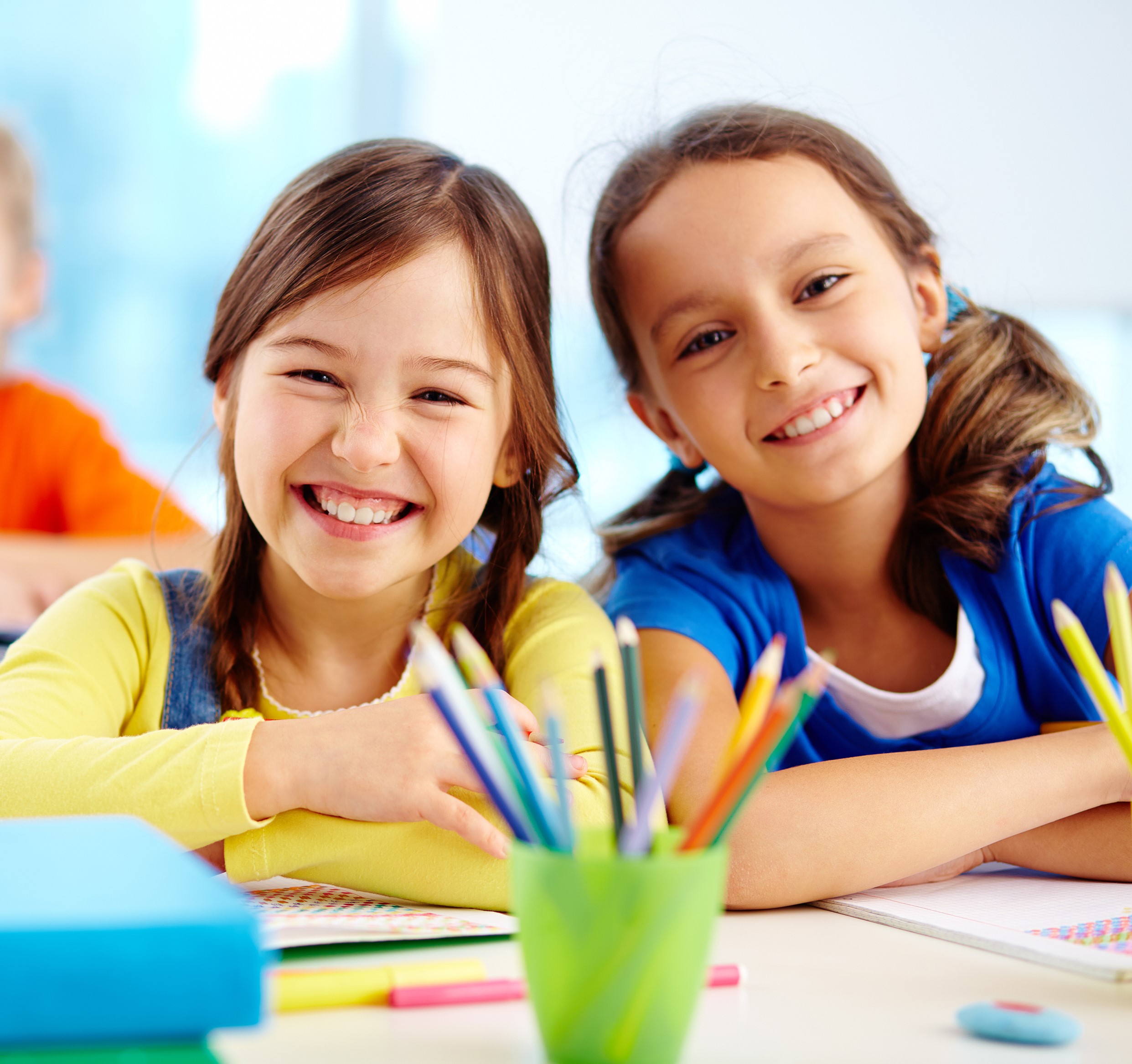 Two young female students sitting at a desk at school with a cup of colored pencils.